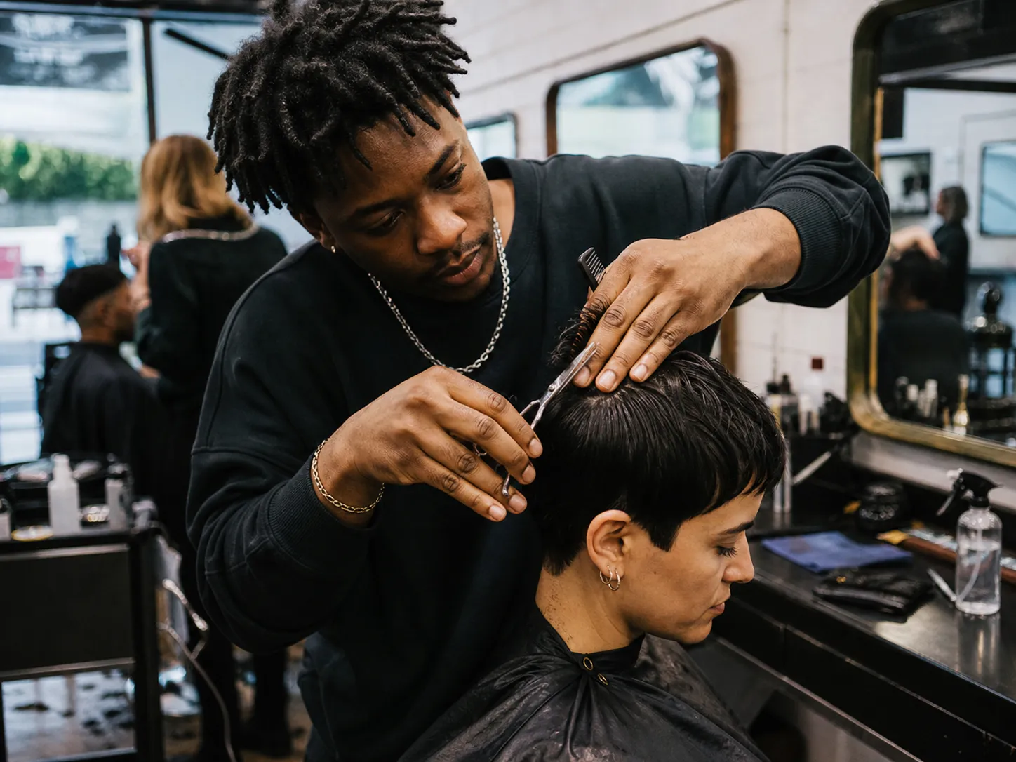 Ein Friseur schneidet konzentriert das kurze Haar einer Kundin in einem belebten Salon.